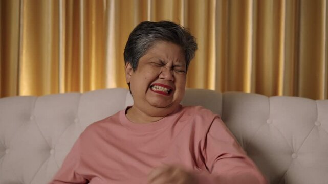 Video footage medium shot, captured face of Asian Senior woman short-haired, Showing signs of irritation, brooding, and dissatisfaction expressing while sitting on sofa in living room