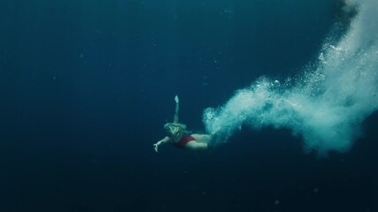 Teen girl in red suit jumps in the water from the boat and swims confidently underwater in the crystal clear tropical sea with sun shining through the water