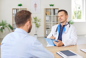 Serious focused male doctor in lab coat sitting at desk with clipboard, carefully listening to...