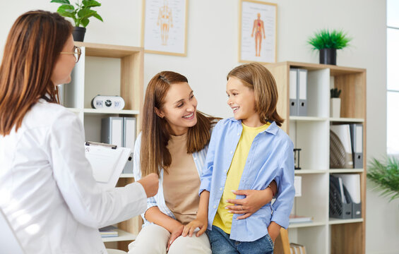 Little smiling girl patient standing near her mother during consultation in medical clinic. Friendly pediatrician in lab coat talking to mother and child, providing health checkup. Children health.