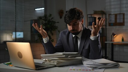 Adult business man manager sitting at desk working on laptop in office in evening, tired expression looking at stack of paper documents holding hands up.