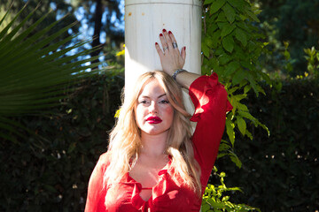 young and beautiful blonde woman, makeup, dressed in red and elegant, rests her hand and back on a...