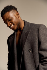 A young handsome African American man poses confidently, showcasing his charm and style with an open coat. The soft lighting highlights his features and joyful demeanor against a neutral backdrop.