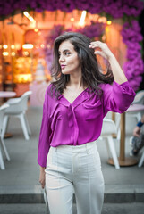 Fototapeta premium Young beautiful brunette woman in a pink jacket and white pants posing on the street. Portrait of a beautiful young woman in a purple blouse and white pants posing in an outdoor cafe on the street