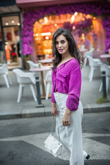 Young beautiful brunette woman in a pink jacket and white pants posing on the street. Portrait of a beautiful young woman in a purple blouse and white pants posing in an outdoor cafe on the street