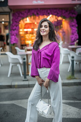 Young beautiful brunette woman in a pink jacket and white pants posing on the street. Portrait of a beautiful young woman in a purple blouse and white pants posing in an outdoor cafe on the street