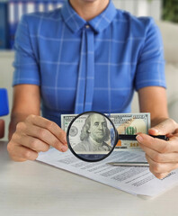 Female banker carefully examines designation on money through magnifying glass on table in office