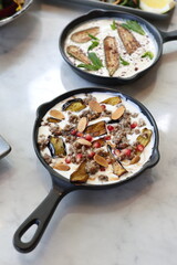 A skillet of Lebanese eggplant fatteh, layered with fried eggplant, creamy yogurt sauce, toasted bread, chickpeas, and garnished with fresh mint and sumac, showcasing a traditional and flavorful Middl