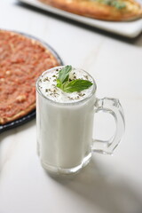 A traditional Lebanese manoushe topped with seasoned minced meat, served on a black tray beside a glass of laban yogurt drink, presented on a marble table.