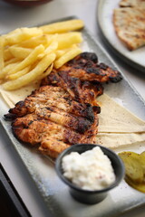 Close-up of a grilled chicken breast, perfectly charred and juicy, served alongside crispy golden French fries. The dish is accompanied by a small black bowl of creamy garlic sauce.