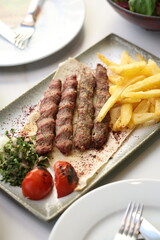 A platter of chargrilled Lebanese beef kebabs served with crispy French fries, grilled tomatoes, onions, fresh parsley, and sumac seasoning on flatbread, offering a hearty and flavorful Middle Eastern