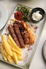 A Lebanese mixed grill platter featuring chargrilled beef kebabs, chicken skewers, and lamb skewers, served with French fries, garlic sauce, grilled tomatoes, fresh parsley, and sumac on flatbread, of