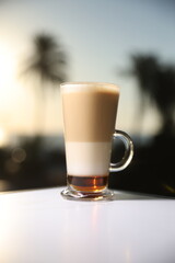 Elegant layered latte in a tall glass, perfectly blending frothy milk and espresso, set against a warm tropical sunset with palm silhouettes.