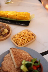 A delightful table setup featuring boiled corn on the cob, lupini beans, sesame flatbread, and a salad bowl with fresh vegetables, offering a wholesome snack platter.