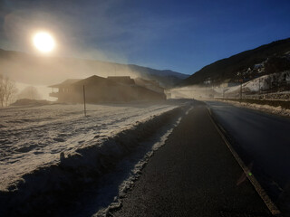 Winter Morning with Foggy Sunlight Over a Snowy Rural Road and Mountain Landscape