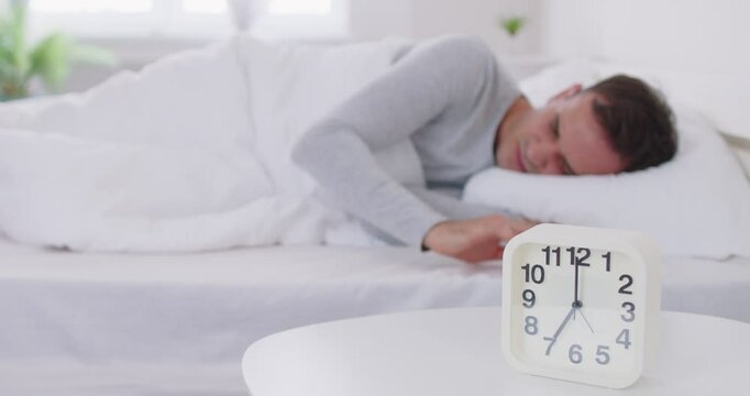 Sleeping man lying on bed in his bedroom and stopping alarm clock at home. Wake up in morning of working day. Young sleepy guy reaching for alarm clock to switch it off. 4k video. Slow motion video.