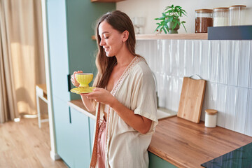 Satisfied joyful attractive smiling girl drinking tea from yellow cup and saucer in scandinavian style kitchen at home