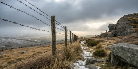 Obraz premium A rugged barbed wire fence in a cold and hard environment with rocky outcrops and sparse vegetation, set against a grey sky with low-hanging clouds, frosty conditions, grey sky