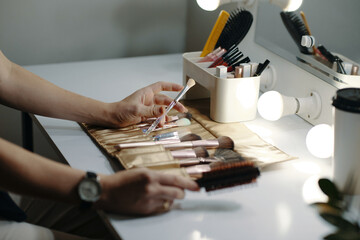 Close-up of hands selecting makeup brushes from a roll-up pouch on a vanity desk with cosmetic...