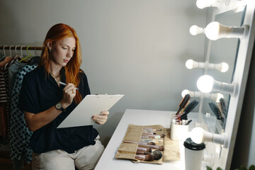 Female makeup artist sitting by illuminated vanity mirror, holding clipboard and examining work....