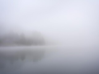 Fototapeta premium Softly falling fog wraps around the lake's edge, veiling the surrounding trees in a misty embrace, gentle mist, atmospheric conditions, still water
