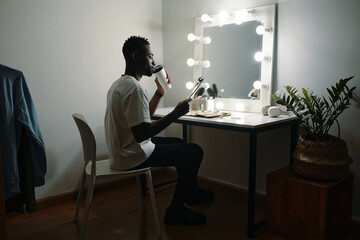 African American man grooming in well-lit dressing room with vanity mirror and plants, holding makeup brush while seated at chic wooden table
