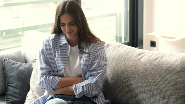 A young woman sits on the sofa, massaging her abdomen to ease the discomfort from acute stomach pain caused by gastroenteritis, stress, constipation, and menstrual cycle- People with stomach ache 