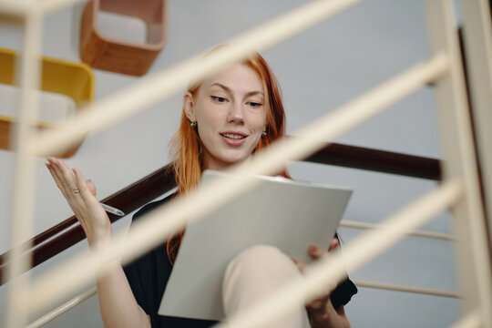 Woman sitting on staircase holding notepad and pen, smiling while writing notes. Surrounded by a modern interior with shelving in background