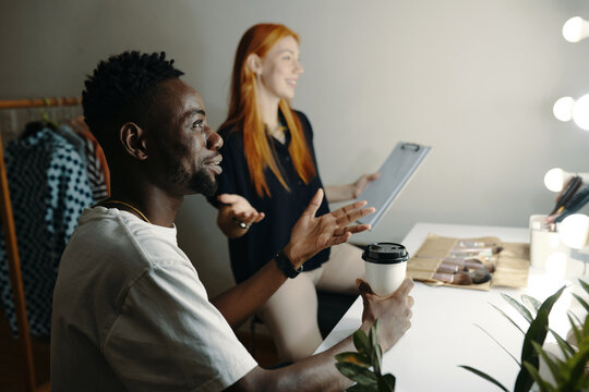 Man holding coffee cup engaging in conversation with woman holding tablet in dressing room with bright lights and clothing racks visible in background
