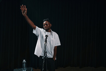 Fototapeta premium Individual standing on stage with a microphone, holding left hand aloft, possibly giving speech or performing with water bottle on stand visible. Dark curtain background emphasizing subject