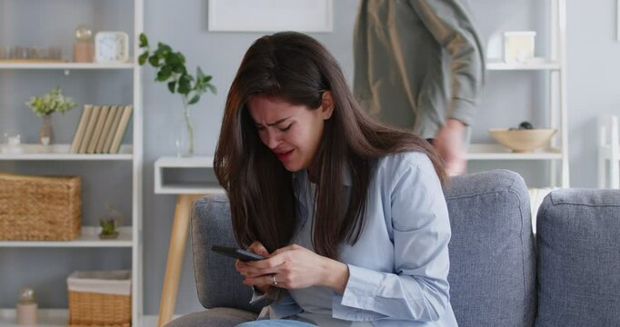 Quarreling young angry man shouting on his upset crying wife using mobile phone writing a message. Unhappy couple arguing in living room at home. Relationship and divorce concept. 4k video.