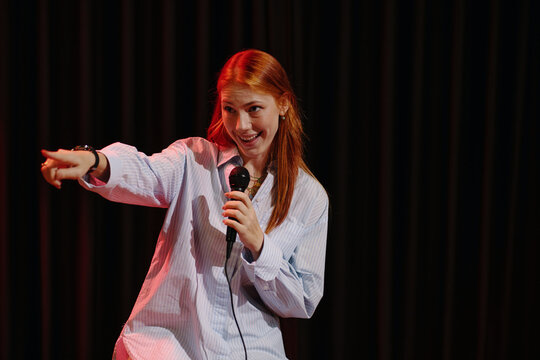 Young Caucasian woman passionately performing stand-up comedy on stage, holding microphone and pointing toward audience. Standing against dark curtain backdrop, engaging crowd with humor and energy