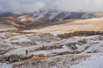 Winter scene in Altai Mountains shows lone hiker on snow-dusted terrain beneath cloudy skies and...
