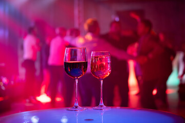 Red and white wine glasses stand on a table while people enjoy a vibrant party with music and dancing blurred in the background.