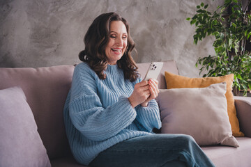 Smiling mature woman relaxes at home in cozy sweater enjoying leisure time with smartphone in bright daylight