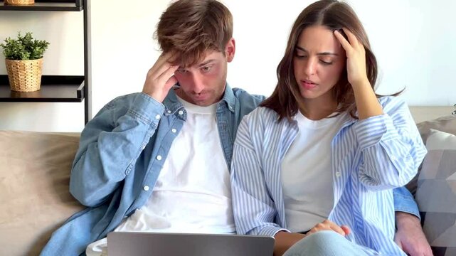 A man and a woman are sitting together on the sofa at home, both looking at a laptop screen with concerned expressions as they read the negative results of an exam or a work-related test