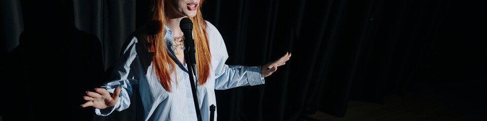 Woman engaged in performing stand-up comedy on stage, holding microphone and gesturing with hands...
