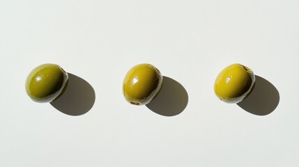 Three fresh green olives positioned in a row, casting soft shadows against a smooth white background, showcasing their glossy surfaces and natural color variations.