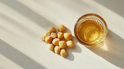 Macadamia oil in a glass jar next to shelled macadamia nuts on a bright surface, viewed from above with soft shadows creating depth.