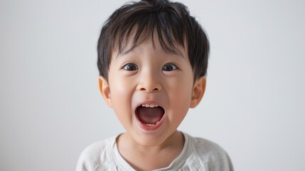 A young boy with dark hair and a white shirt standing in front of a plain background with his mouth open in a wide smile.