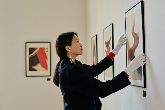 Woman adjusting framed artwork on gallery wall wearing gloves. Modern abstract paintings hanging alongside for exhibition