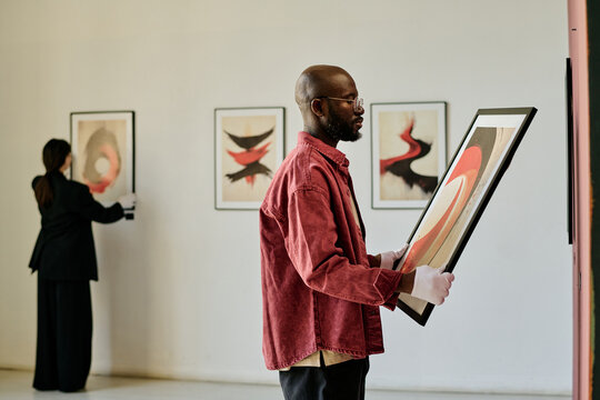 African American man holding abstract painting, curating art gallery exhibit. Background reveals another person examining framed artwork on white walls