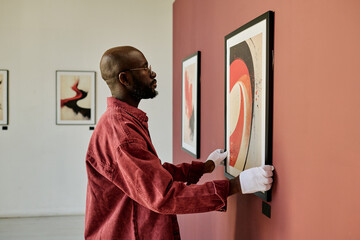 African American man examining abstract artwork hanging on wall in art gallery setting displaying white gloves and red shirt