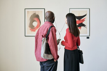Two individuals in an art gallery observing framed paintings on a white wall while engaged in...