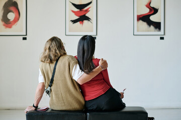 Two women sitting on black bench, admiring abstract paintings on gallery wall, arm around each...