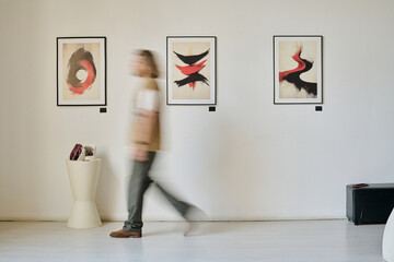 Blurred figure walking through art gallery exhibiting abstract paintings on wall demonstrating...