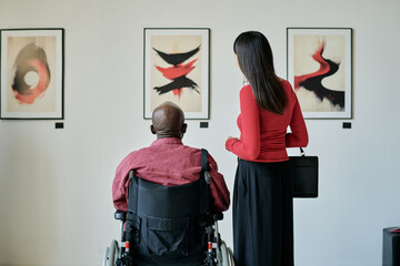 Two people observing abstract artworks displayed on wall in modern art gallery. One person in...