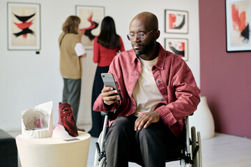 Man in wheelchair looking at smartphone while visiting an art exhibit, with artwork and people...