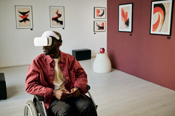 Man in wheelchair exploring modern art gallery using virtual reality headset with abstract paintings on walls and contemporary decor elements surrounding him