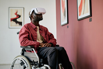 African American man sitting in wheelchair using VR headset in modern art gallery. Engaging with virtual reality technology displayed in frame